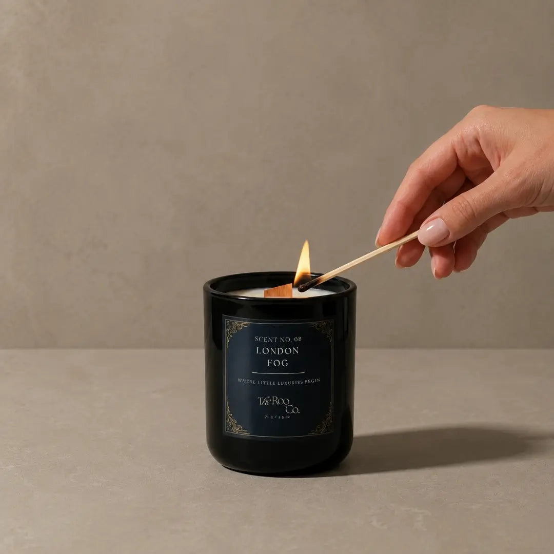 Luxury 2.5 oz black candle with a wooden wick and navy label on a soft neutral plaster background. London Fog scented candle inspired by Earl Grey tea, bergamot, vanilla bean, burnt sugar, and creamy milk notes is lit with a wooden match held by a person's hand, set against a neutral textured beige background.