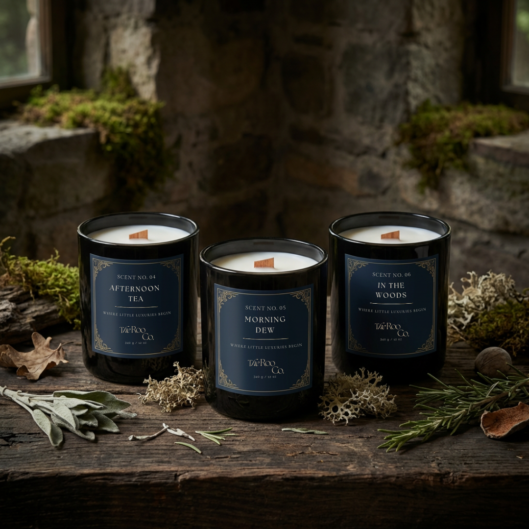 Aromatic candle collection by The Roo Co including Afternoon Tea, Morning Dew, and In the Woods, displayed on rustic wood with herbs and greenery in a natural, greenhouse-inspired setting.