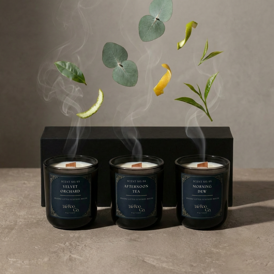 Three candles with labels 'Velvet Orchard', 'Afternoon Tea', and 'Morning Dew' on a gray surface with floating leaves and smoke.