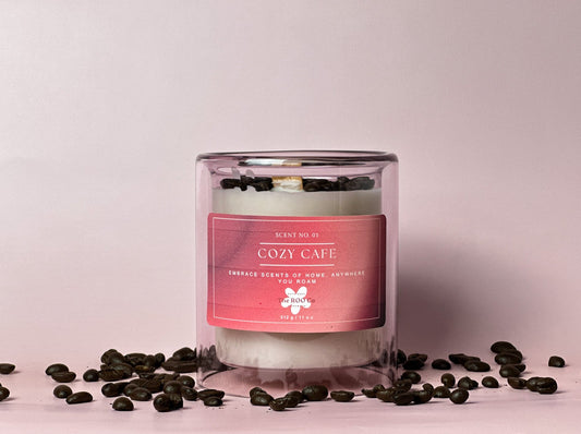 A luxury candle called cozy cafe that is 11 oz in a light violet vessel. In the background there is a cup of coffee, a jar of sugar, and spilled coffee beans. Great for gifts for any occasion.