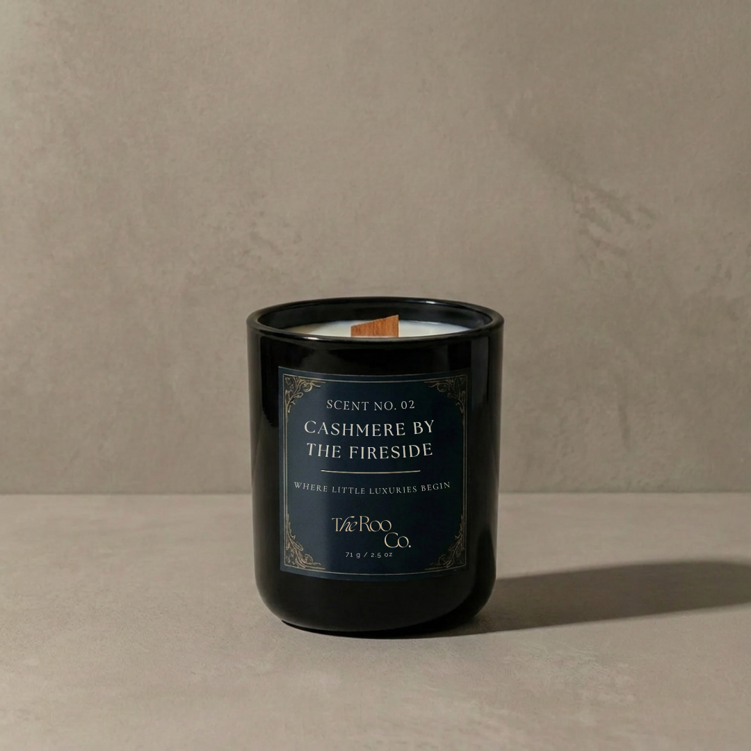  Luxury 2.5 oz black candle with a wooden wick and navy label on a soft neutral plaster background. Cashmere by the Fireside scented candle inspired by warm woods, soft cashmere, amber, and smoky fireside notes.