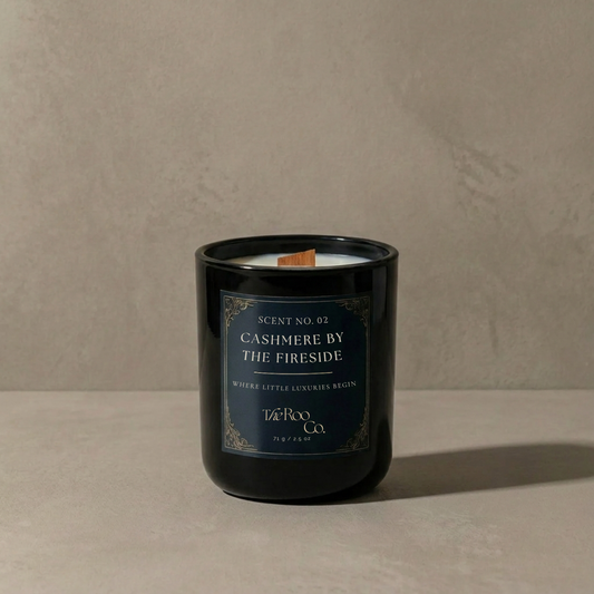  Luxury 2.5 oz black candle with a wooden wick and navy label on a soft neutral plaster background. Cashmere by the Fireside scented candle inspired by warm woods, soft cashmere, amber, and smoky fireside notes.