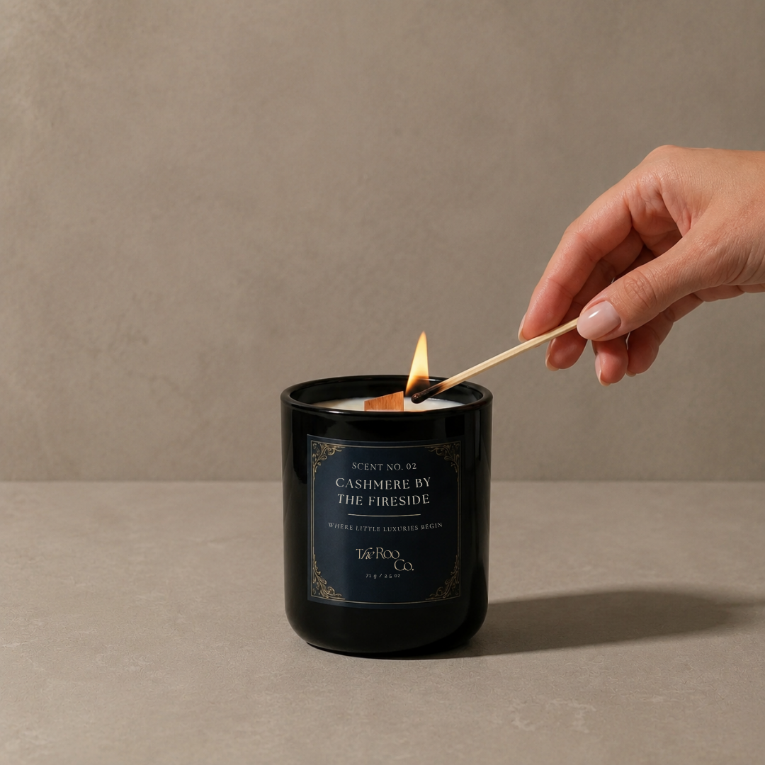 cashmere by the fireside luxury coconut soy candle in a glossy black glass jar with a wooden wick on a neutral beige background with hand lighting a candle