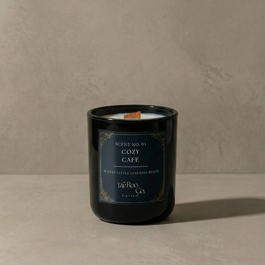 Luxury 2.5 oz black candle with a wooden wick and navy label on a soft neutral plaster background.     Cozy Café scented candle inspired by coffee, cacao, caramelized sugar, and creamy vanilla notes.